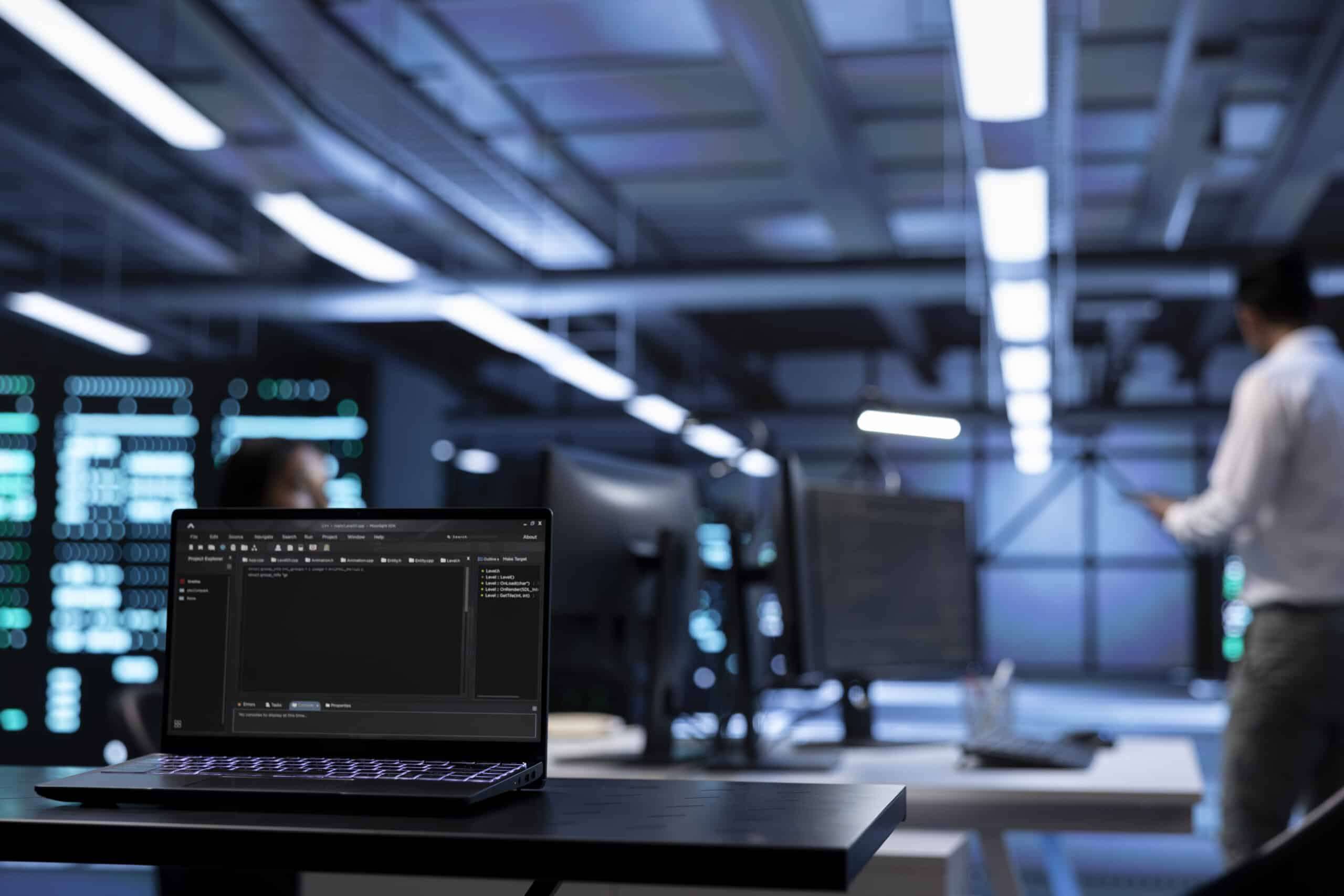 A laptop showing an IDE on a desk in a dark, modern office, with a colleague at a screen wall in the background
