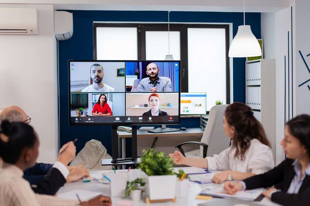 Varios compañeros alrededor de la mesa de una sala de reuniones miran una pantalla de pared con cuatro personas en videollamada