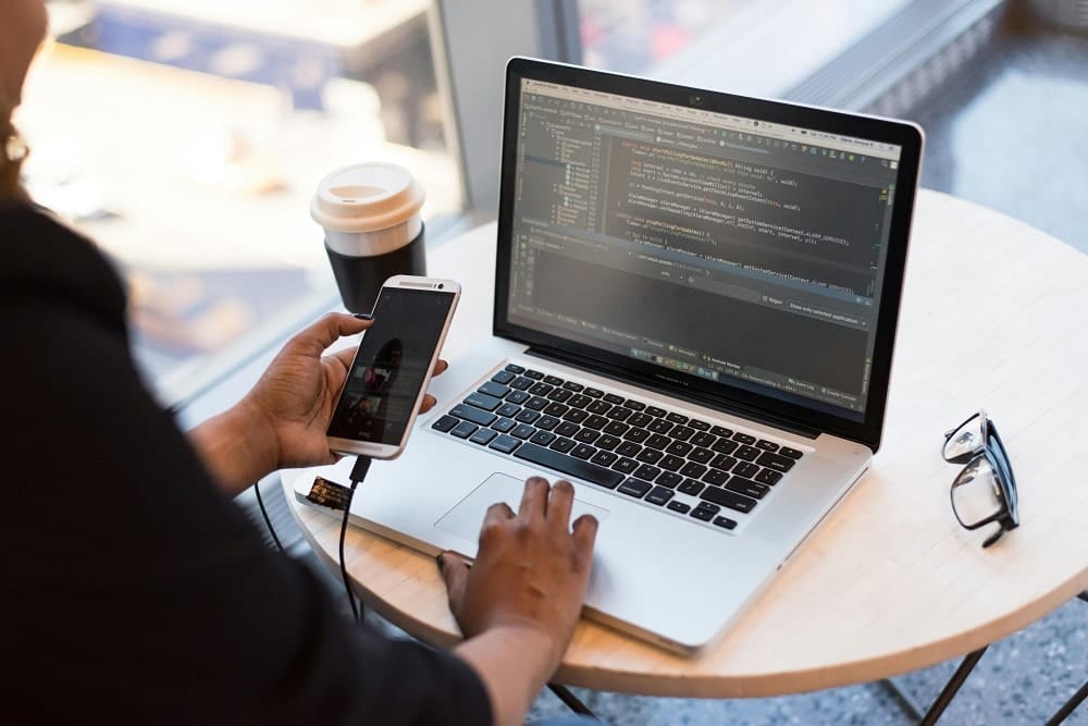 A person holding a phone in one hand while typing on a laptop that shows an IDE, with a coffee cup and glasses on the table