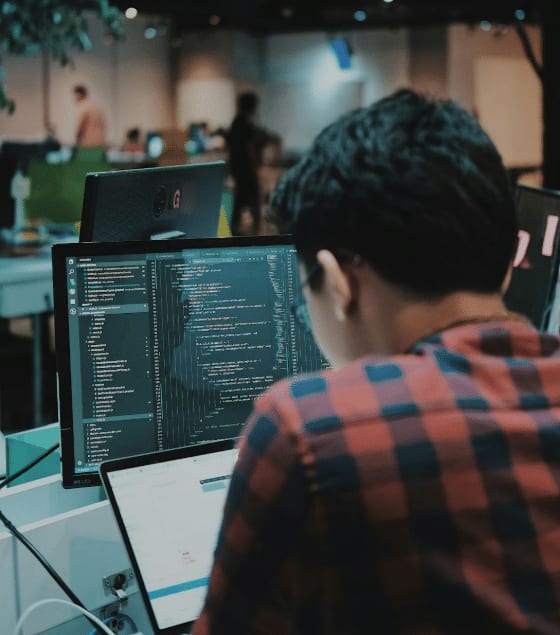 Una persona desarrolladora vista de espaldas trabaja ante un monitor lleno de código en una oficina diáfana
