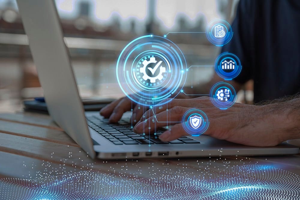 Hands typing on a laptop with a glowing tick-in-a-gear icon and linked quality, analytics and shield icons above the keyboard