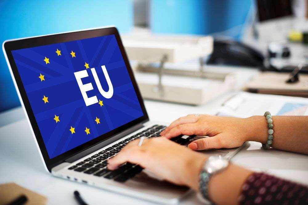 Unas manos teclean en un portátil cuya pantalla muestra la bandera de la Unión Europea y las letras «EU»