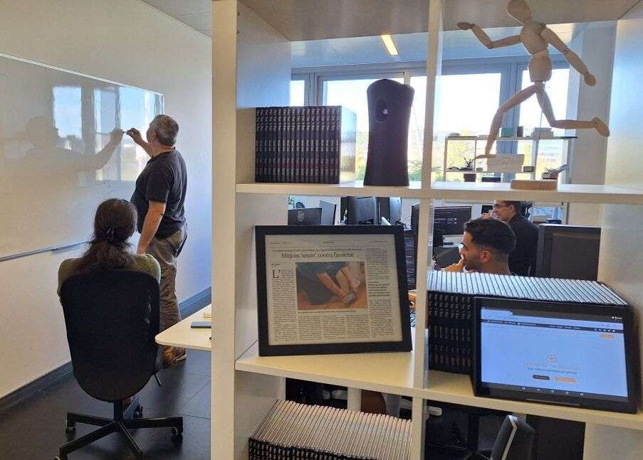 Dues persones desenvolupadores treballant a les seves pantalles a l’oficina d’Unimedia mentre una companya escriu en una pissarra