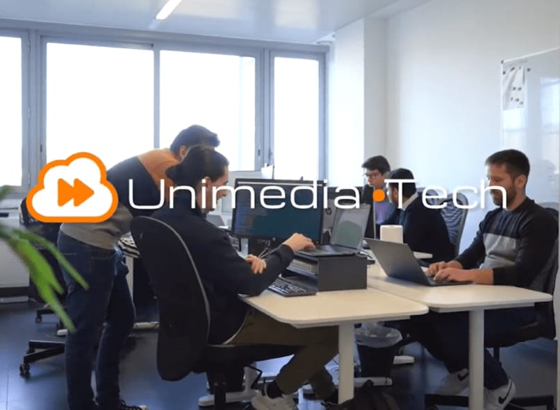 El logotipo de Unimedia Technology sobre una fotografía del equipo trabajando en sus mesas en una oficina luminosa