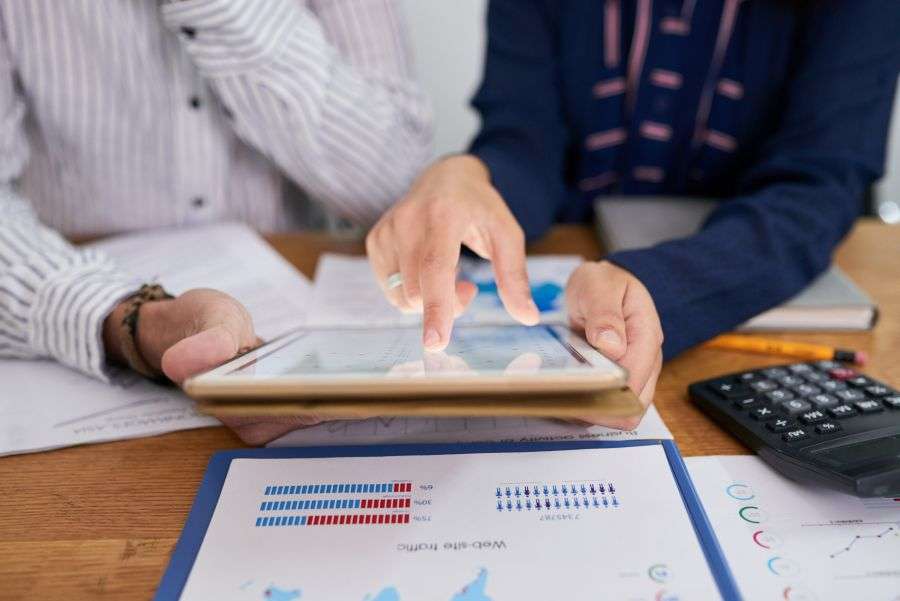 Two colleagues reviewing figures on a tablet across a desk covered in printed charts, beside a calculator